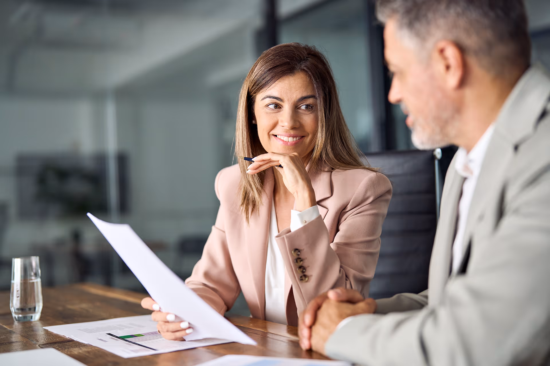 Une femme souriante en veste rose tenant un document, discutant avec un homme en costume gris dans un bureau.
