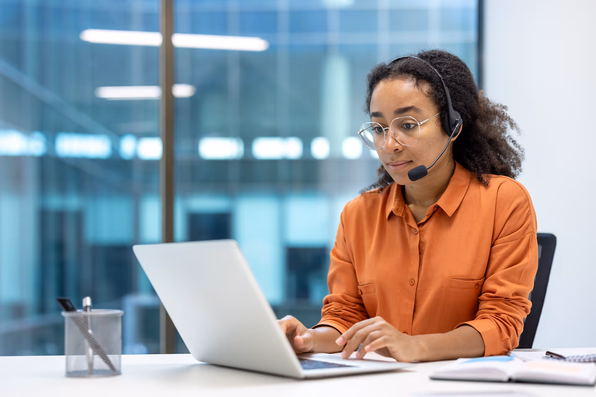 Femme portant un casque-micro et des lunettes, travaillant sur un ordinateur portable au bureau.