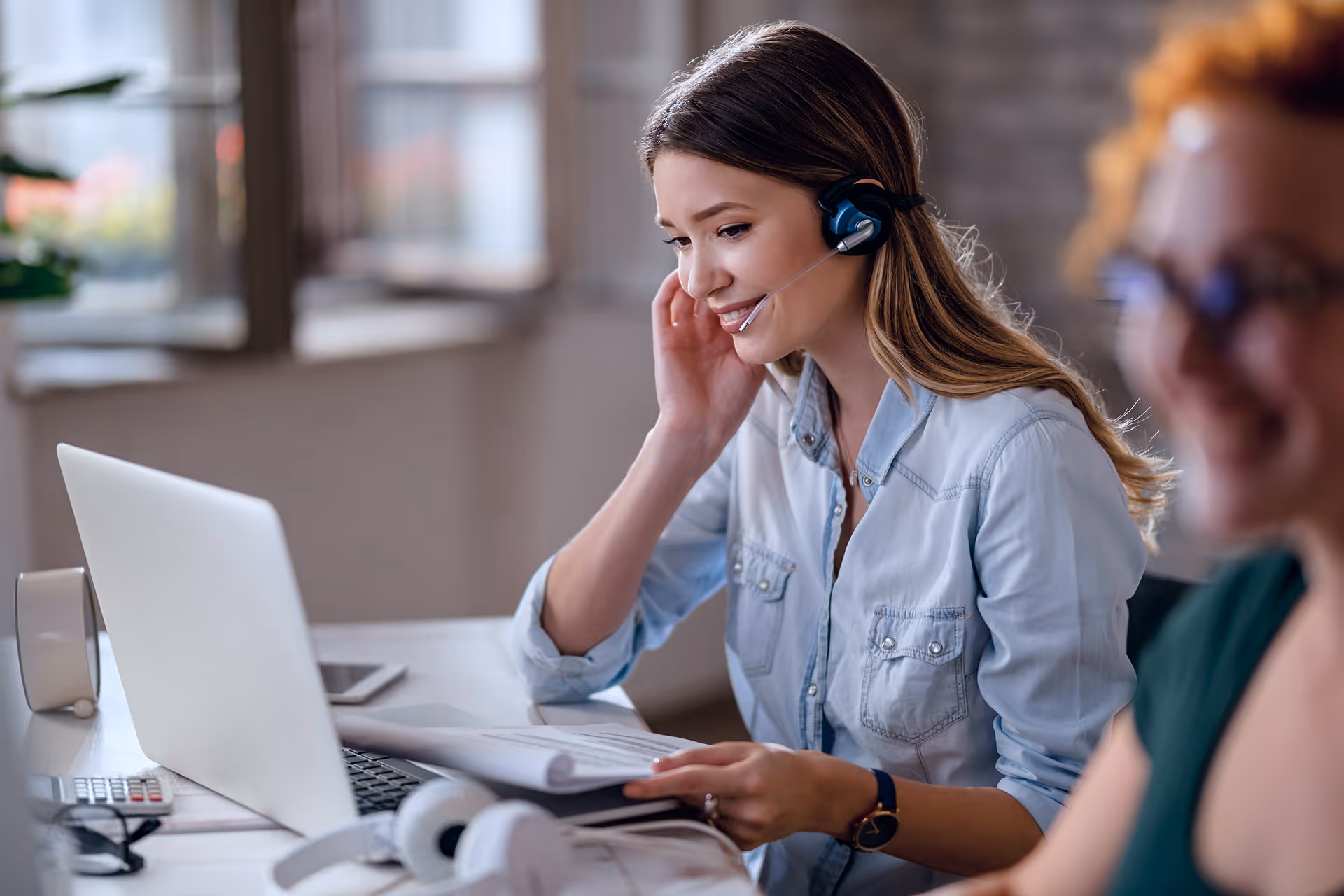 Femme souriante portant un casque avec microphone, travaillant devant un ordinateur portable et consultant des documents.