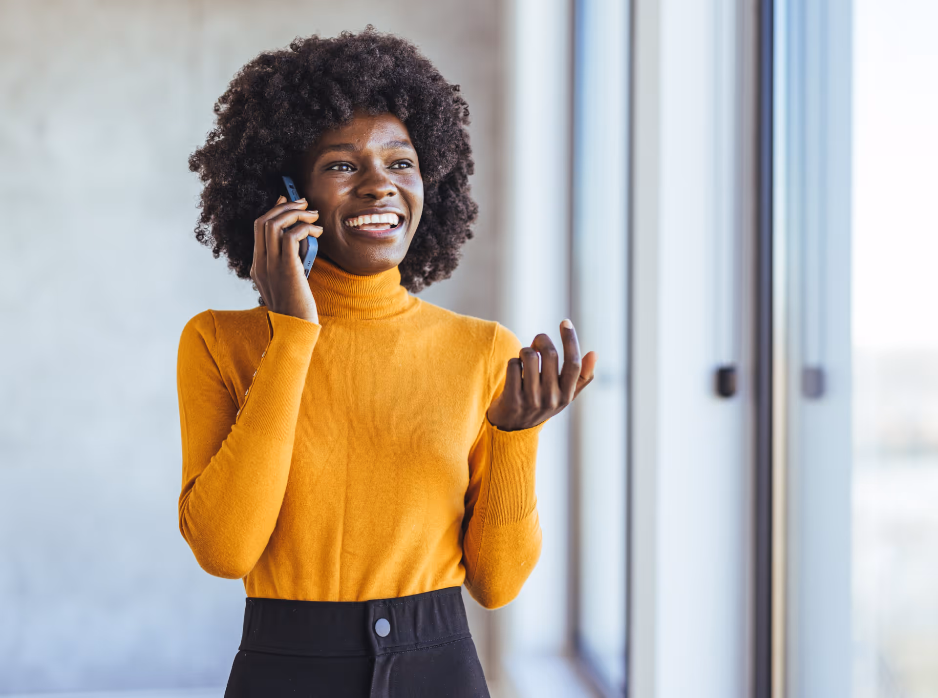 Femme souriante portant un chandail à col roulé orange parlant au téléphone près d'une fenêtre.
