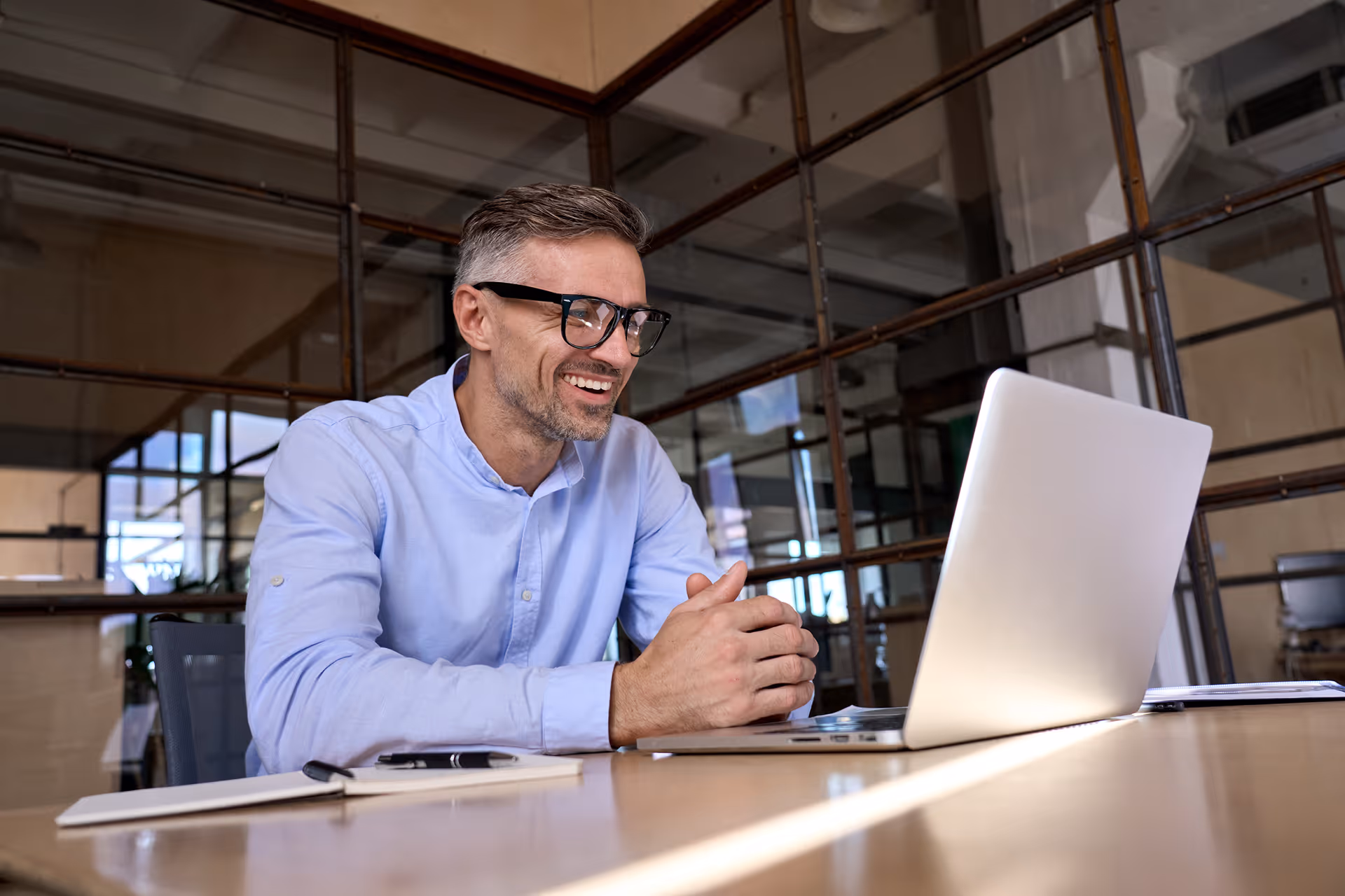 Homme souriant en chemise bleue portant des lunettes, assis à une table avec un ordinateur portable dans un bureau moderne.