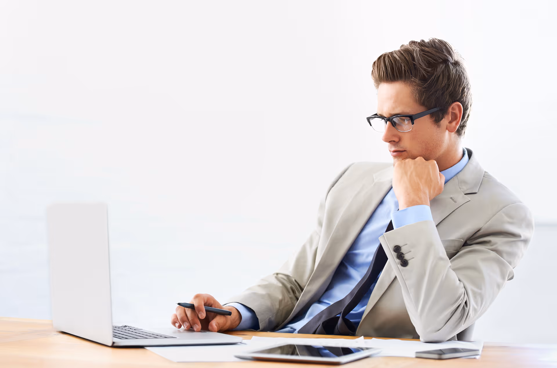 Homme d'affaires portant des lunettes et un costume beige, assis à un bureau en réfléchissant devant un ordinateur portable.