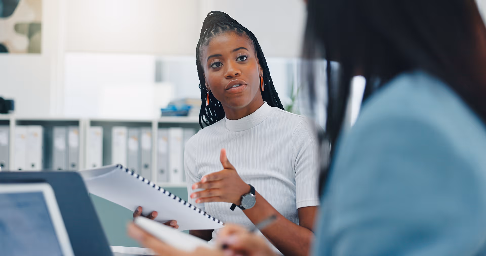 Jeune femme noire parlant lors d'une réunion, tenant un cahier à spirale, avec une autre personne en premier plan floue.