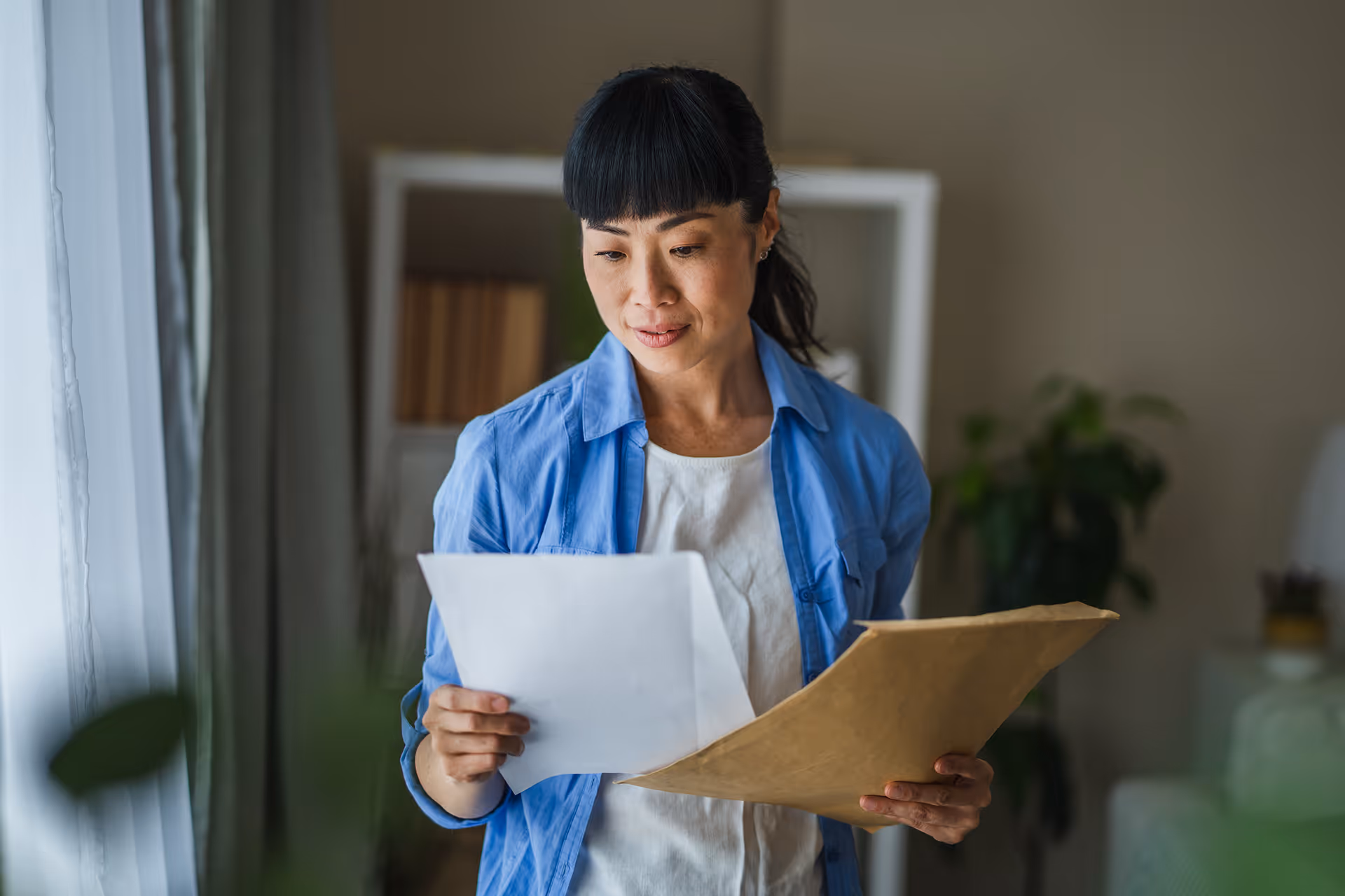 Femme asiatique tenant des papiers, lisant attentivement à l'intérieur d'une maison.