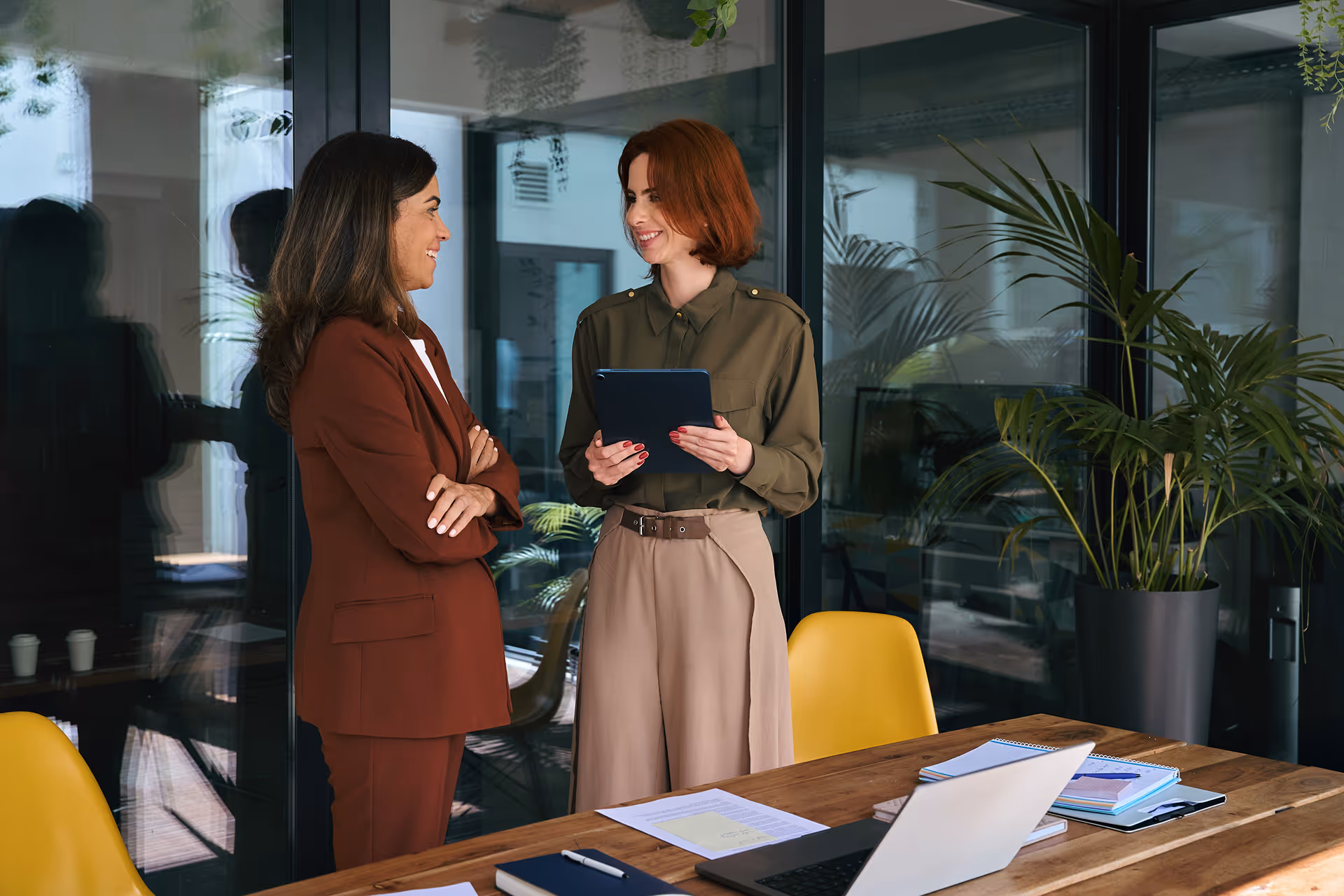 Deux femmes collègues discutant souriantes dans un bureau moderne avec tablette, ordinateur portable et plantes.