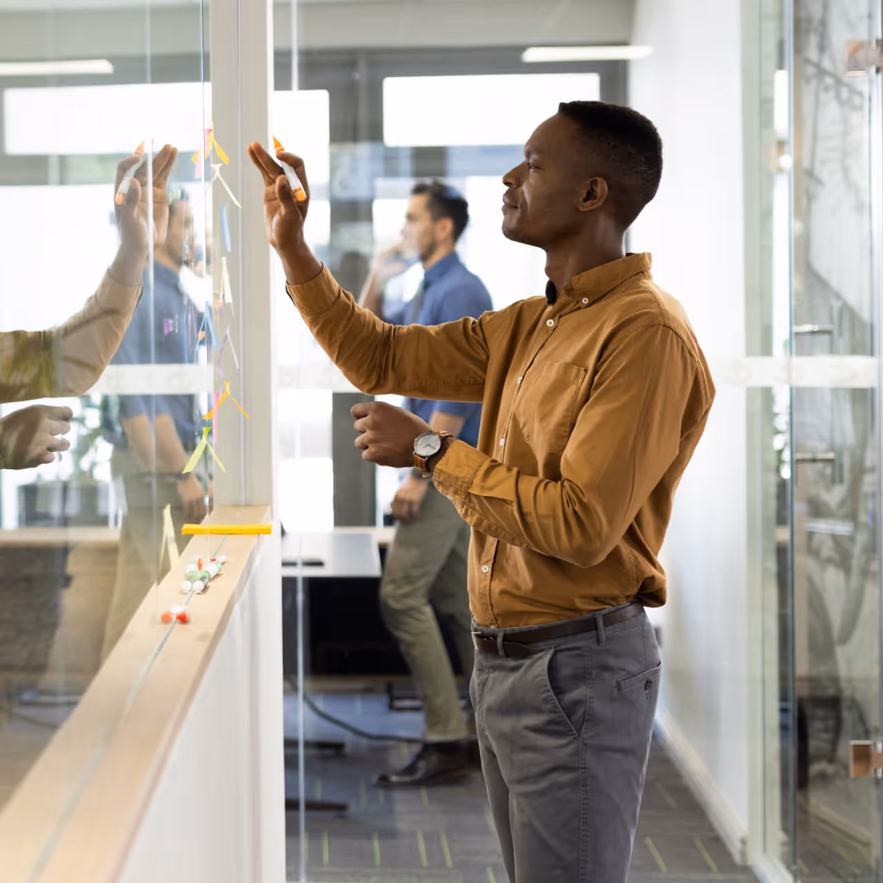 Un homme écrit sur un tableau en verre avec un marqueur orange, un autre homme est visible à l'arrière-plan en train de parler au téléphone dans un bureau moderne.