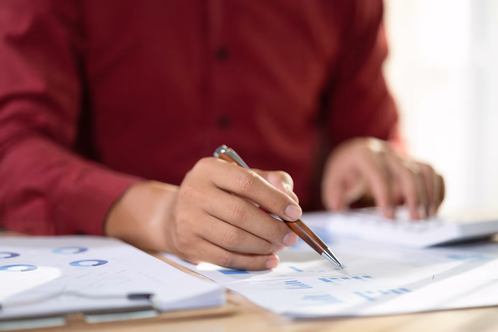 Une personne en chemise rouge analyse des graphiques financiers avec un stylo à la main sur un bureau.