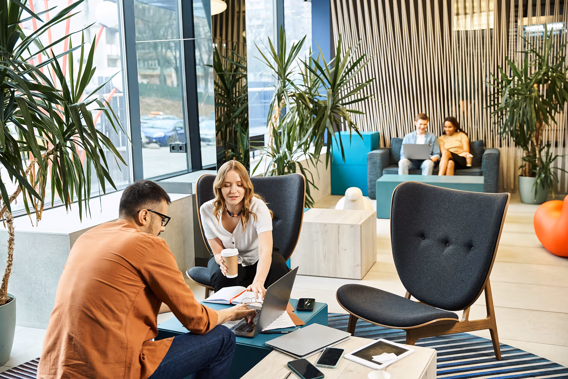 Deux paires de collègues collaborant dans un espace de travail moderne avec des plantes vertes et des fauteuils confortables.