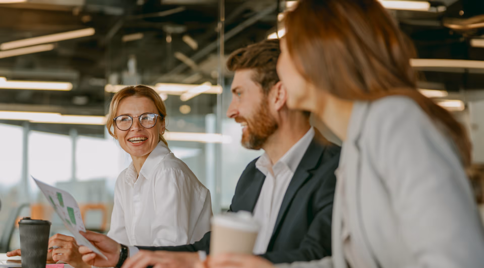 Trois collègues souriant et discutant lors d'une réunion en bureau moderne, une femme tenant des documents.