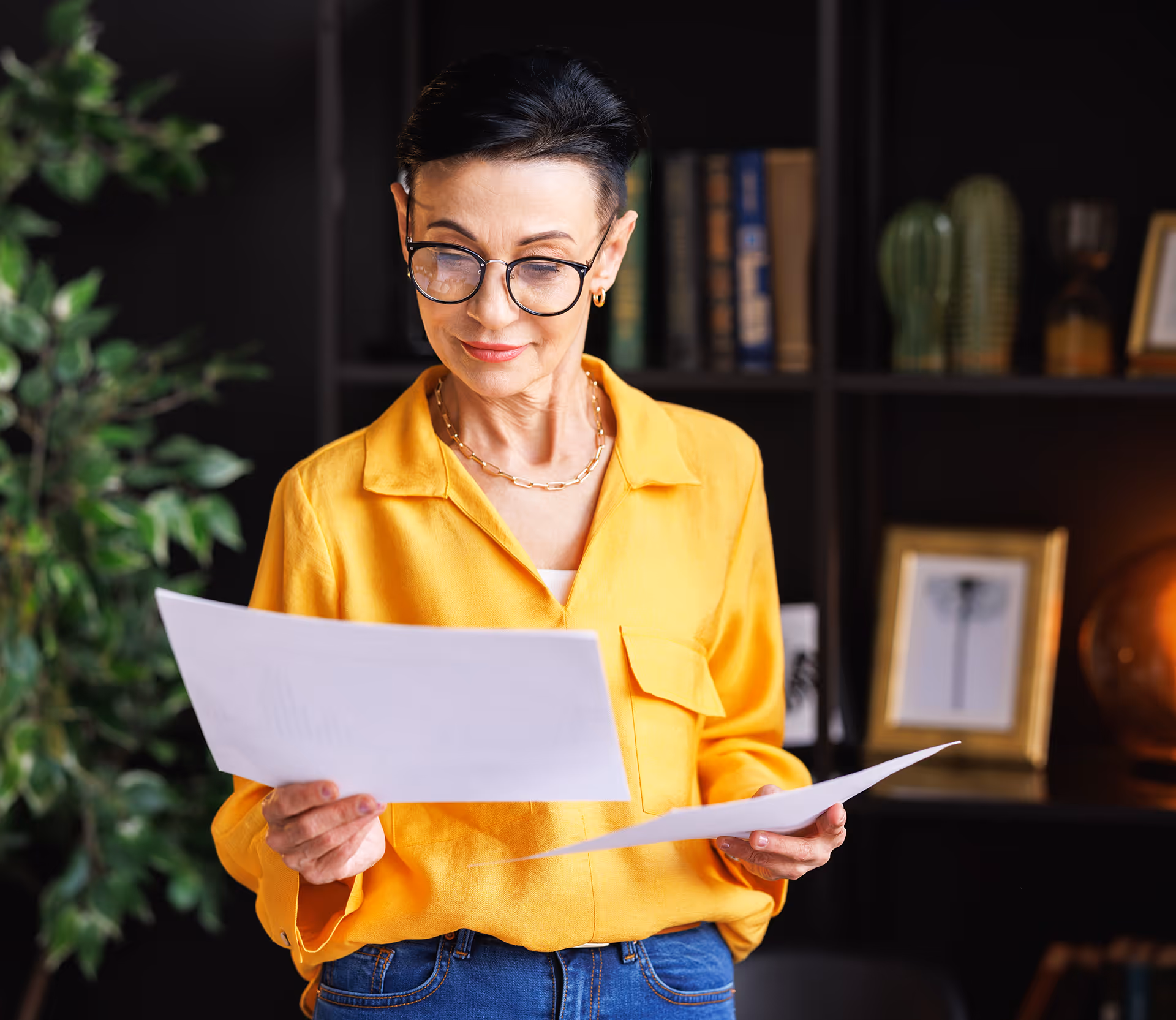 Femme portant des lunettes et une chemise jaune lisant des documents dans une pièce avec une bibliothèque et des plantes en arrière-plan.