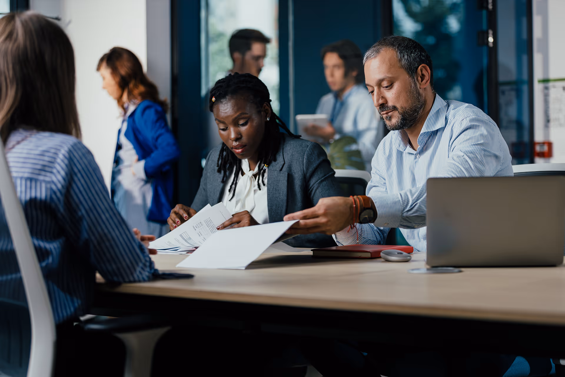 Deux collègues d’affaires assis à une table de réunion examinent des documents avec un ordinateur portable devant eux, tandis que d’autres personnes discutent en arrière-plan dans un bureau moderne.