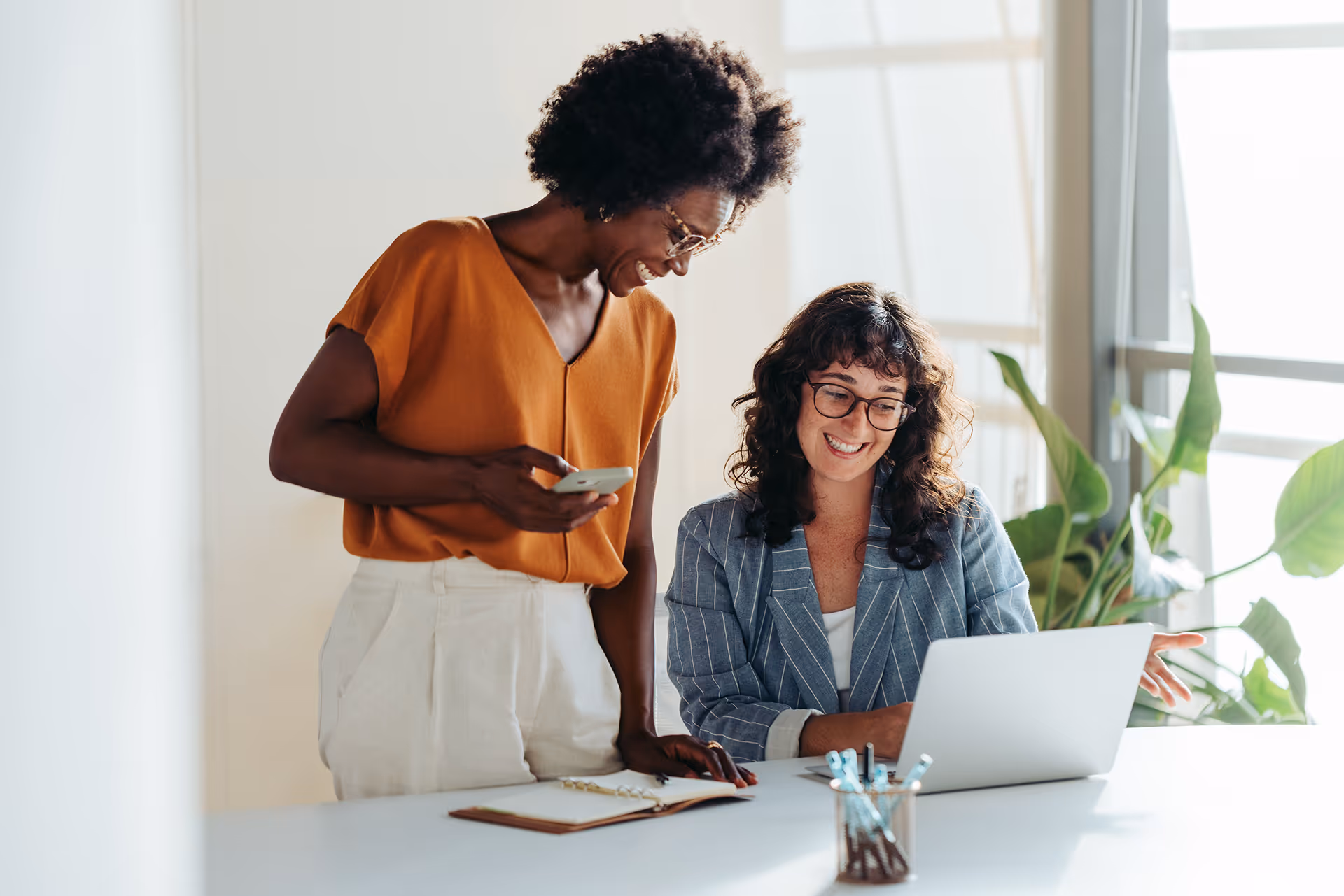 Deux collègues souriantes, l'une debout avec un téléphone et l'autre assise devant un ordinateur portable dans un bureau lumineux avec des plantes.