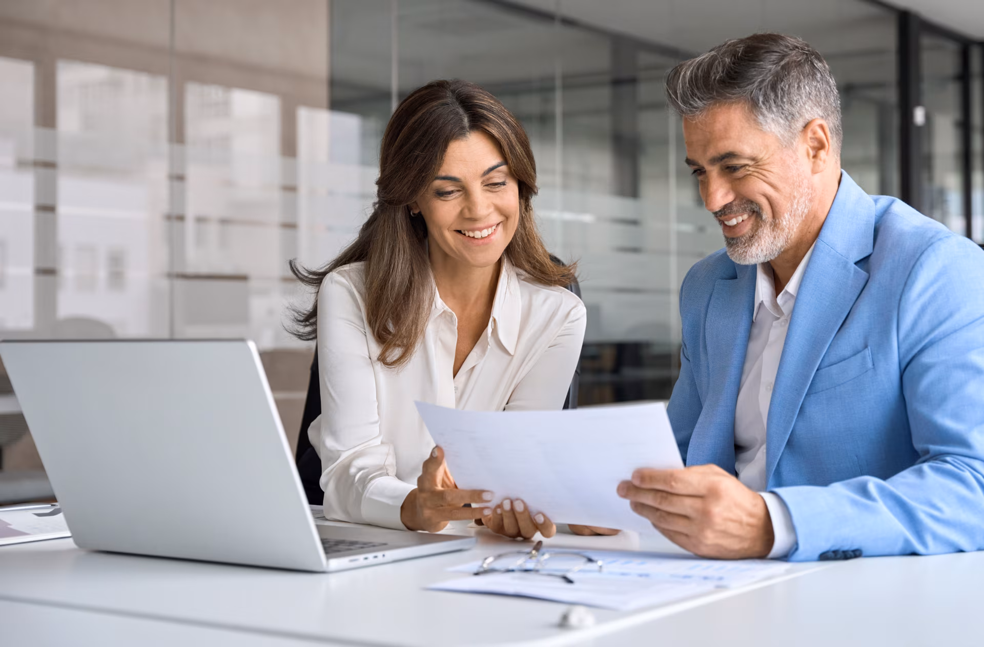 Deux collègues souriants discutent autour d'un document dans un bureau moderne avec un ordinateur portable.