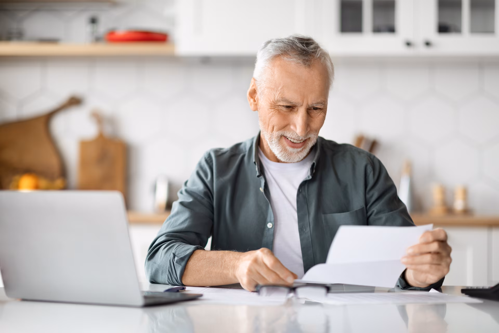 Homme âgé souriant lisant un document à sa table avec un ordinateur portable devant lui dans une cuisine moderne.